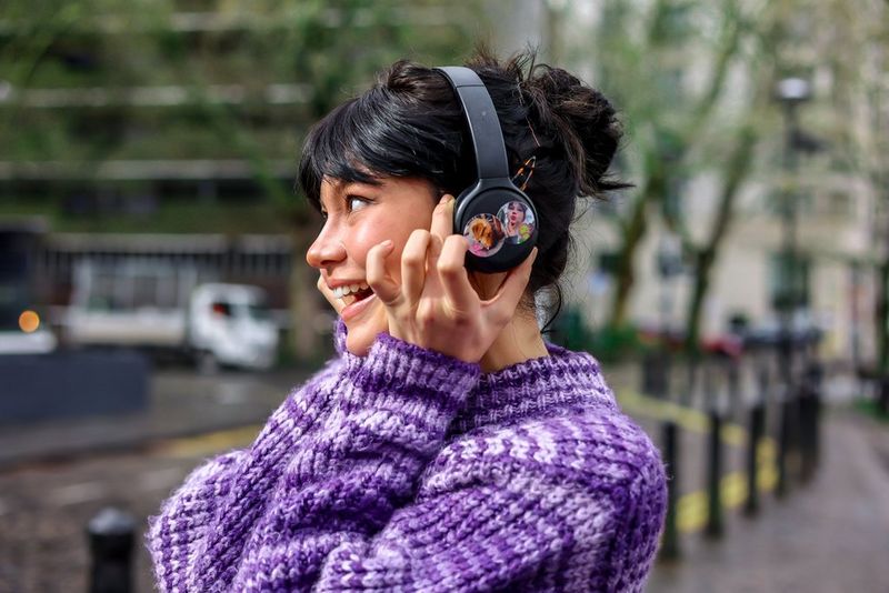 A woman in a purple jumper smiles as she places her hands on a pair of headphones on her head, circular photo stickers brightening up the ear cups.