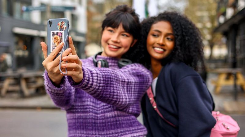 A woman in a purple jumper holds a smartphone to take a selfie while a woman in a dark blue jumper smiles next to her. 