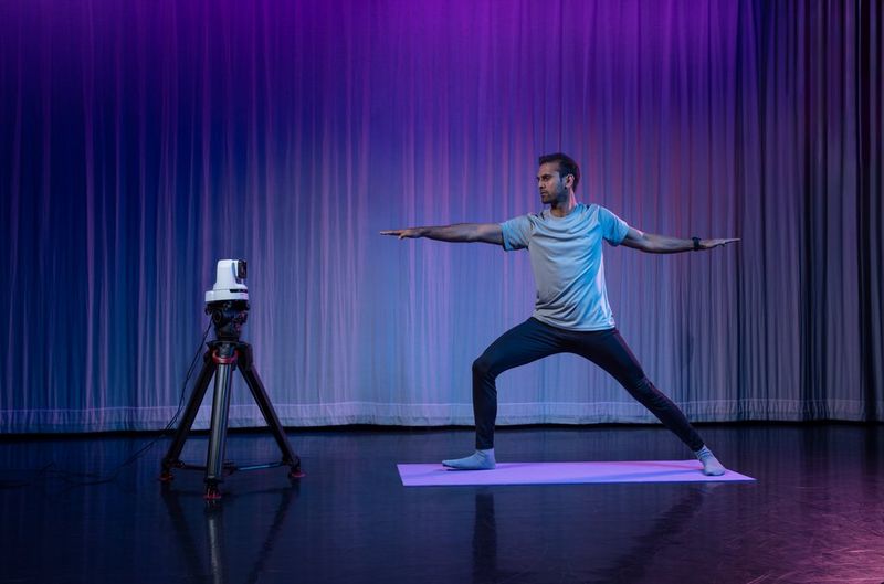 A Canon CR-N100 PTZ camera, mounted on a tripod, points at a man stretching while standing on a yoga mat.