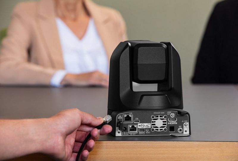A hand reaches out to plug a cable into the back of a Canon CR-N100 PTZ camera.