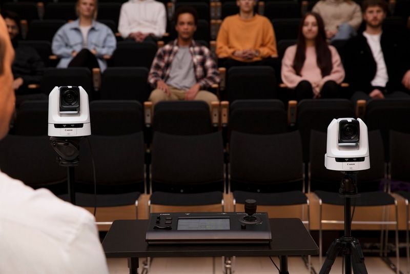 Two Canon CR-N300 cameras stand on tripods in front of the audience in an auditorium, with a Canon RC-IP100 remote camera controller on a table between them.