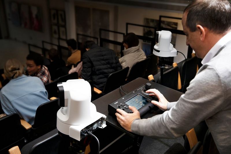 An operator using the RC-IP100 remote camera controller, with two Canon PTZ cameras at the sides of the table at the back of an auditorium.