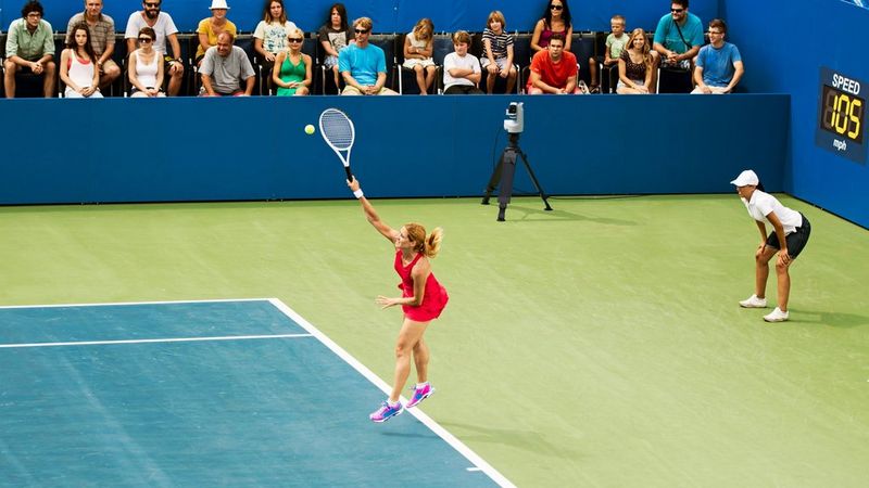 A Canon CR-X300 PTZ camera on a tripod at one side of a tennis court records a player serving.
