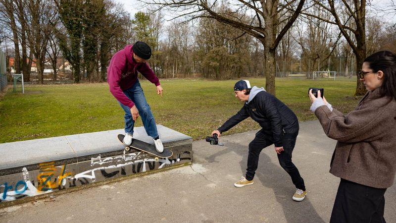 Action photographer Lorenz Holder holds a Canon EOS R5 Mark II with a fisheye lens close to the feet of a skateboarder who is performing a trick in a park. Another photographer stands to one side, shooting from a more conventional angle.