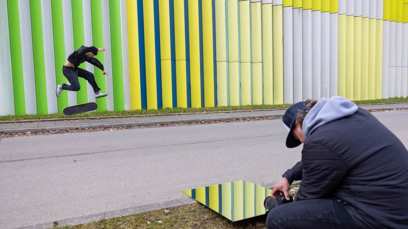 Action sports photographer Lorenz Holder holds his Canon EOS R5 Mark II against a mirror on the ground to photograph the reflection of a skateboarder performing a trick in front of a wall with green and yellow vertical stripes.
