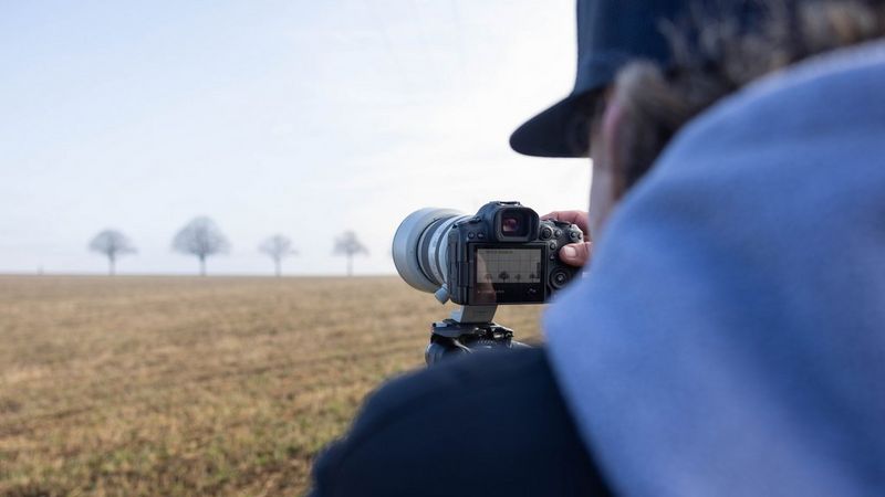 Лоренц Холдер настраивает свою Canon EOS R5 Mark II среди поля, фотографируя скейтбордиста и деревья вдалеке.