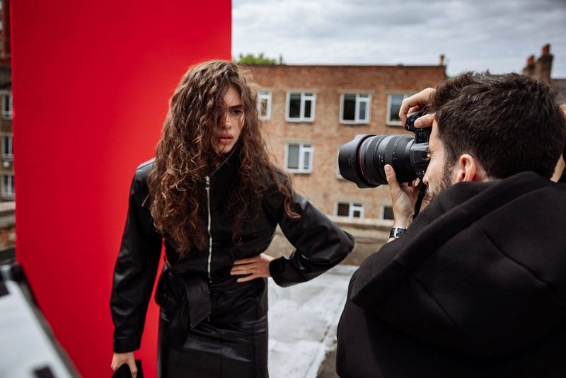 Portrait photographer Samuel Sarfati photographs a model in a dark overcoat in front of a red backdrop on an urban rooftop. 