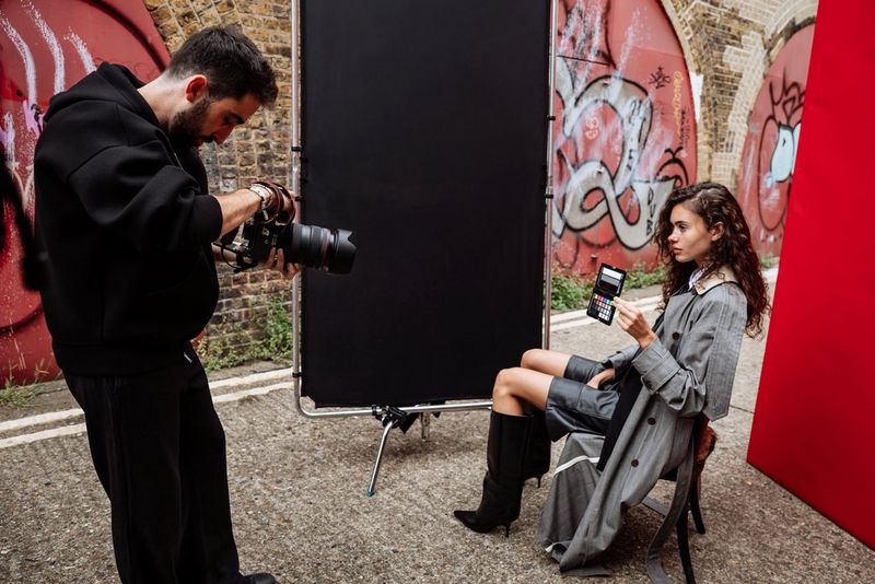 Portrait photographer Samuel Sarfati points his Canon camera at a model, holding a colour reference card as she sits in a narrow urban street setting with graffiti on the wall behind a black negative fill screen.