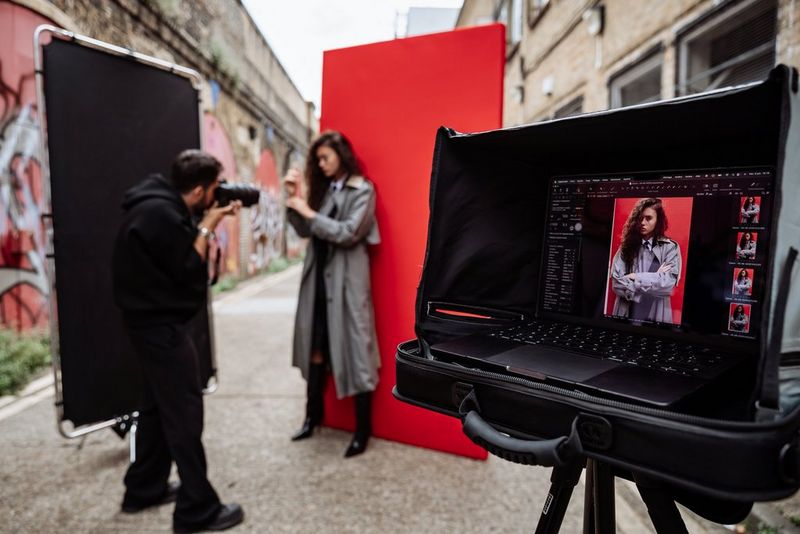 Photographer Samuel Sarfati photographs a model standing in front of a red screen in a narrow urban lane, with images displayed in Capture One on a laptop in the foreground.