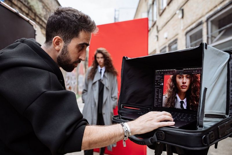 Portrait photographer Samuel Sarfati checks a photo in Capture One on a laptop on a stand in a narrow urban street. A model in a long macintosh stands in front of a red backdrop in the background.