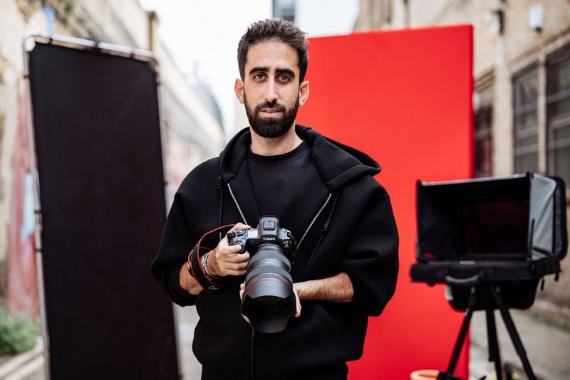 Portrait photographer Samuel Sarfati holds a Canon EOS R5 Mark II with a Canon RF 28-70mm F2L USM lens attached. In the background are a red backdrop and a black light screen.