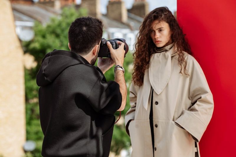 Portrait photographer Samuel Sarfati photographs a model wearing a light-coloured macintosh and standing in front of a red backdrop, with terraced houses out of focus in the background.