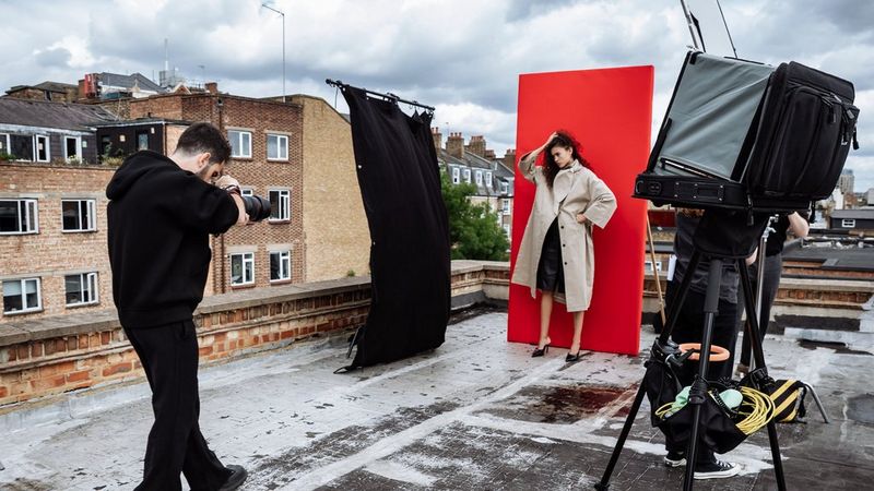 Portrait photographer Samuel Sarfati photographs a model in a cream overcoat standing in front of a red backdrop on a rooftop, with a light box on one side and a black light screen on the other.