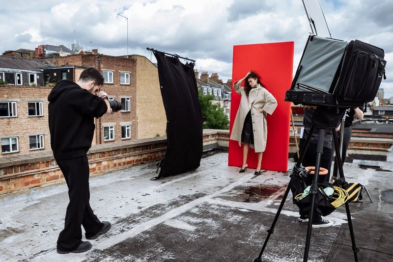 Portrait photographer Samuel Sarfati photographs a model in a cream overcoat standing in front of a red backdrop on a rooftop, with a light box on one side and a black negative fill screen on the other.