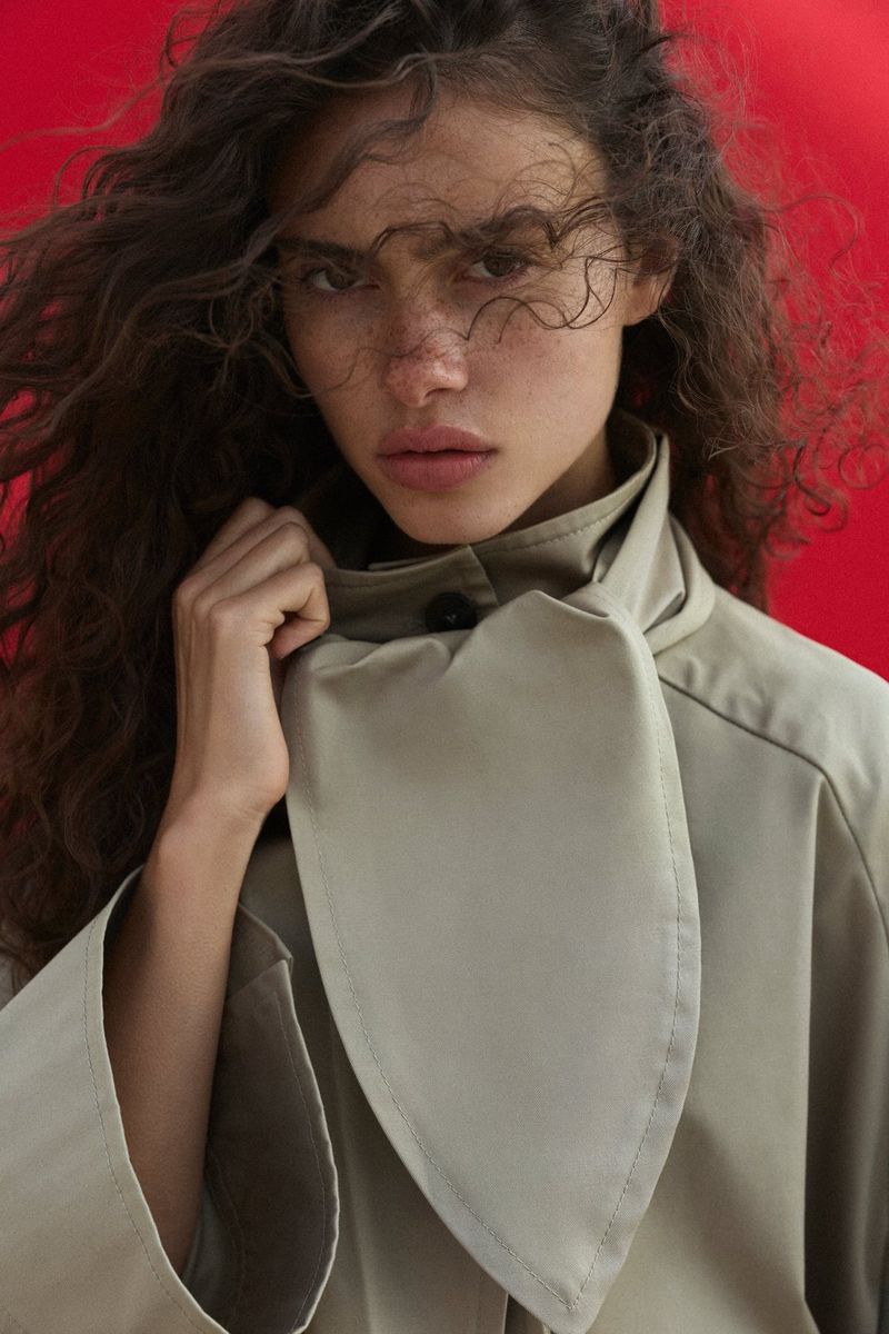 Close-up portrait of a model wearing a light-coloured macintosh in front of a red backdrop, with the wind blowing some of her hair across her face. Natural light portrait photo taken by Samuel Sarfati on a Canon EOS R5 Mark II.
