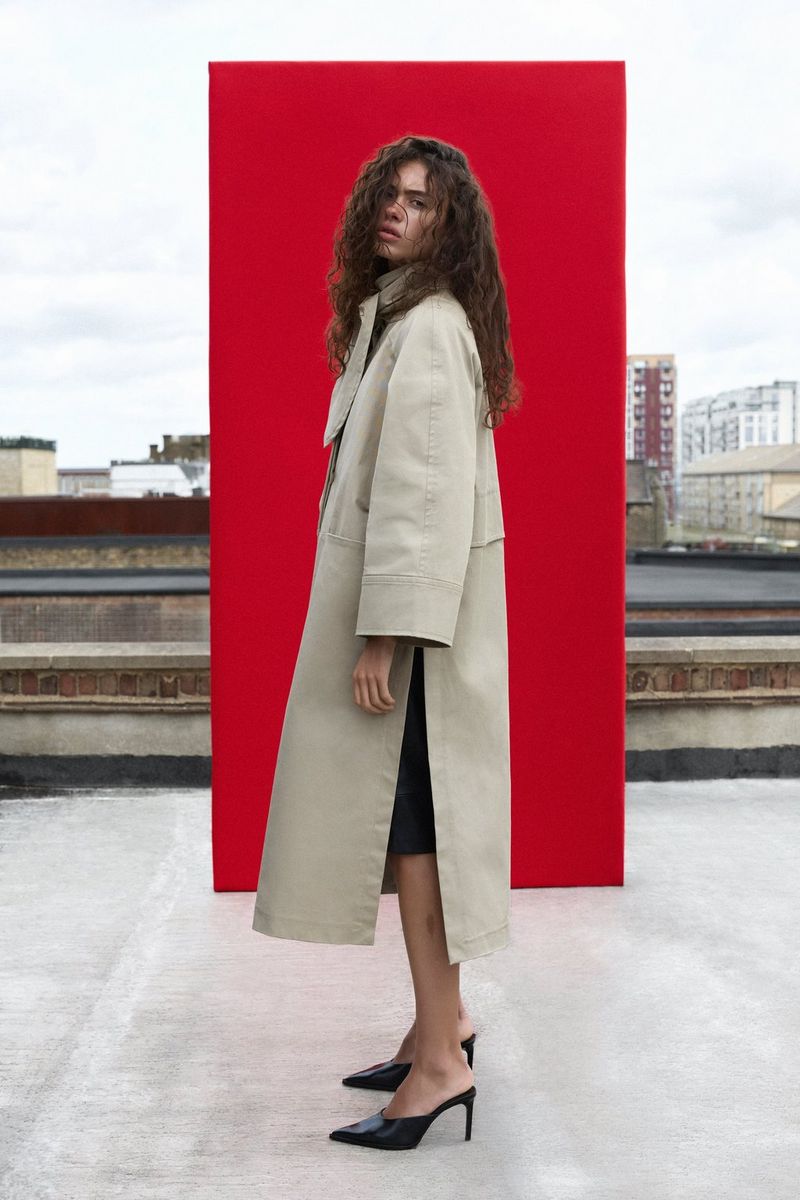 A model wearing a light-coloured macintosh stands in front of a red backdrop on a rooftop, with city roofs visible in the background. Natural light portrait photo taken by Samuel Sarfati on a Canon EOS R5 Mark II.
