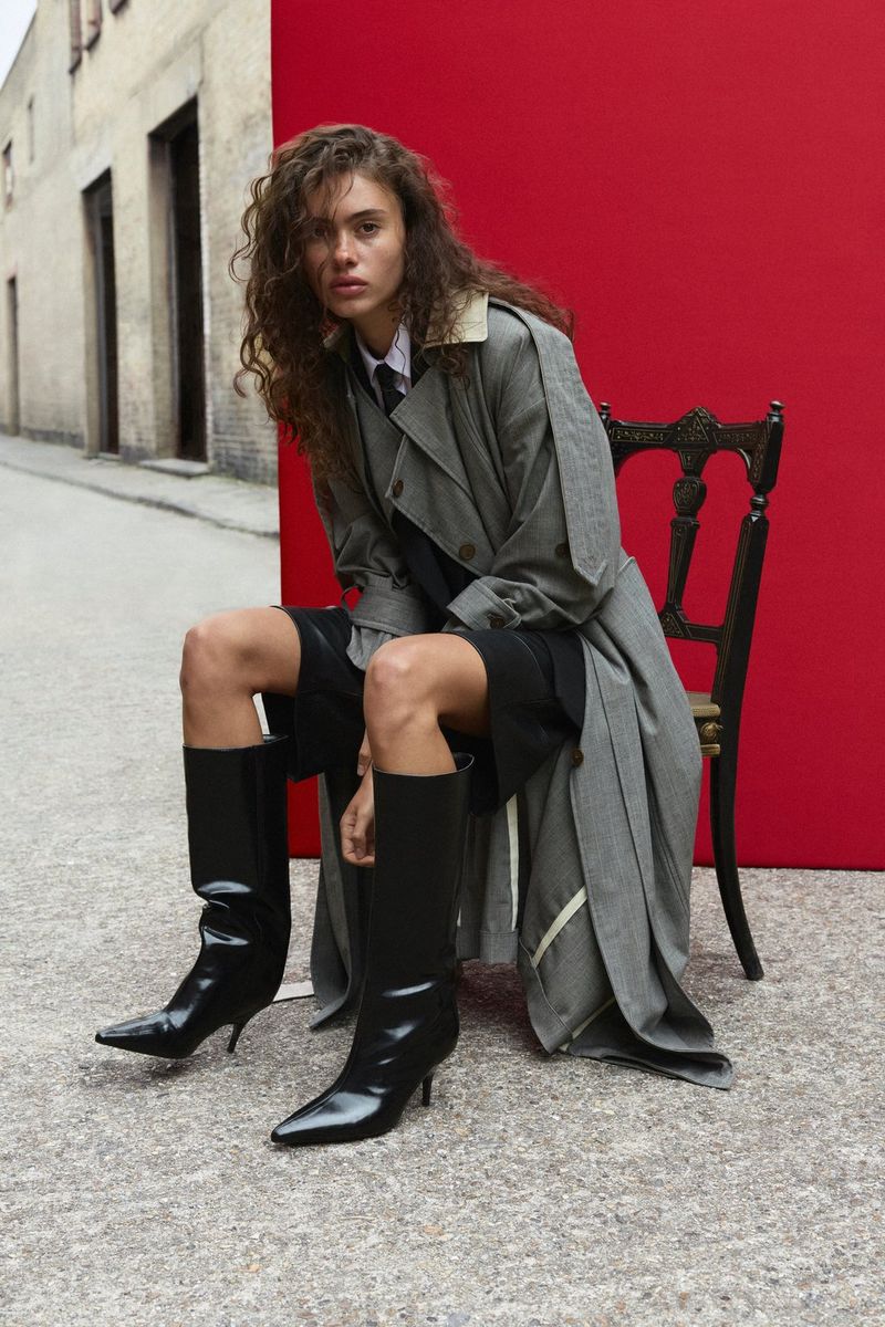 A model wearing a long macintosh and black calf boots sits in front of a red backdrop in a narrow, cobbled urban street. Natural light portrait photo taken by Samuel Sarfati on a Canon EOS R5 Mark II.