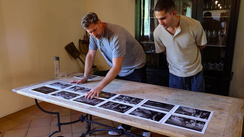 A photographer engages in a portfolio review session with Brent Stirton, examining printed images laid out on a table. 