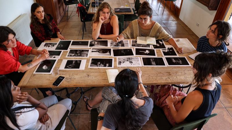 A diverse group of photographers engages in a collaborative portfolio review with Elisa Iannacone, discussing printed photographs laid out on a table.