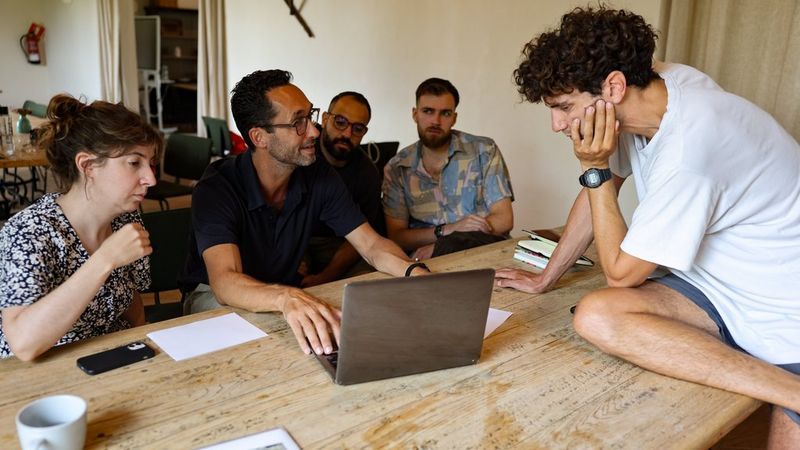Thomas Borberg conducts a portfolio review with a group of photographers, gathered around a laptop at a wooden table at the 2024 Canon Student Development Programme. 