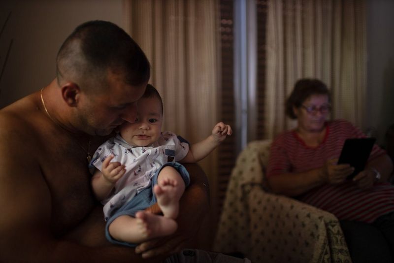A man holding a baby while a woman reads in an armchair in the background. Shot on Canon.
