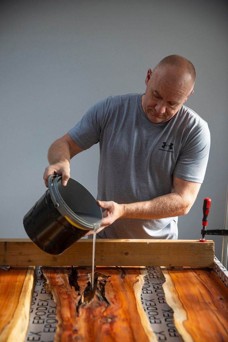 A man pours resin from a bucket into a sheet of reclaimed wood.