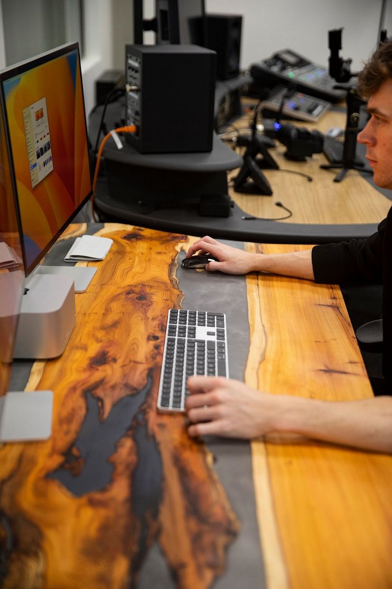 A young man sits at a reclaimed wood table, his hand rests on a Bluetooth keyboard and he looks at a computer screen.