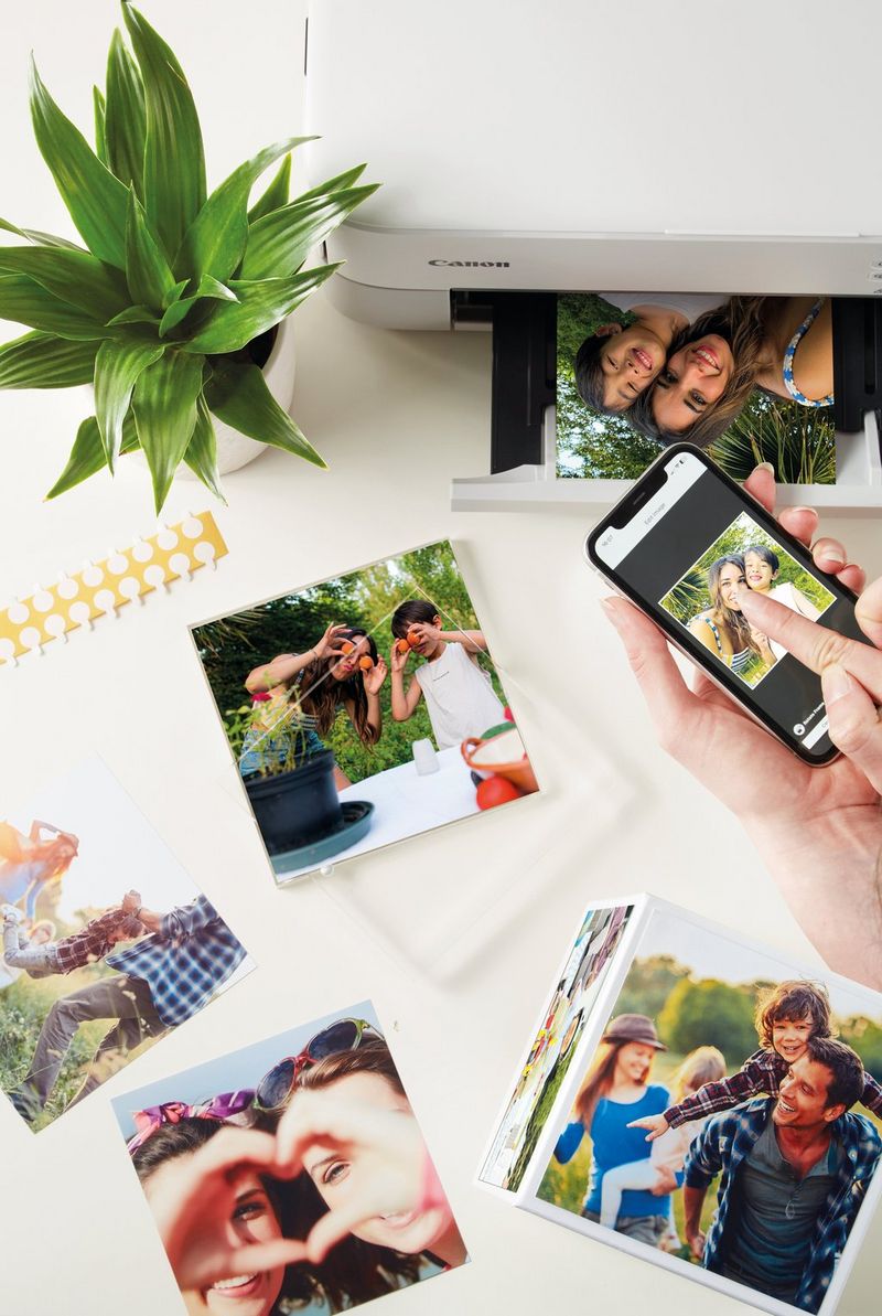 Printed photos lie next to a Canon printer, while a hand holds a phone displaying a photo onscreen. 