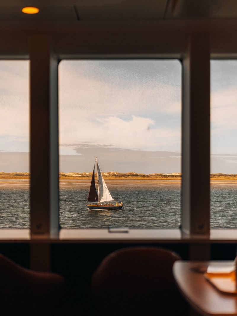 A sailing boat on the water, framed through a window, with blurred chairs and a table just visible in the foreground.