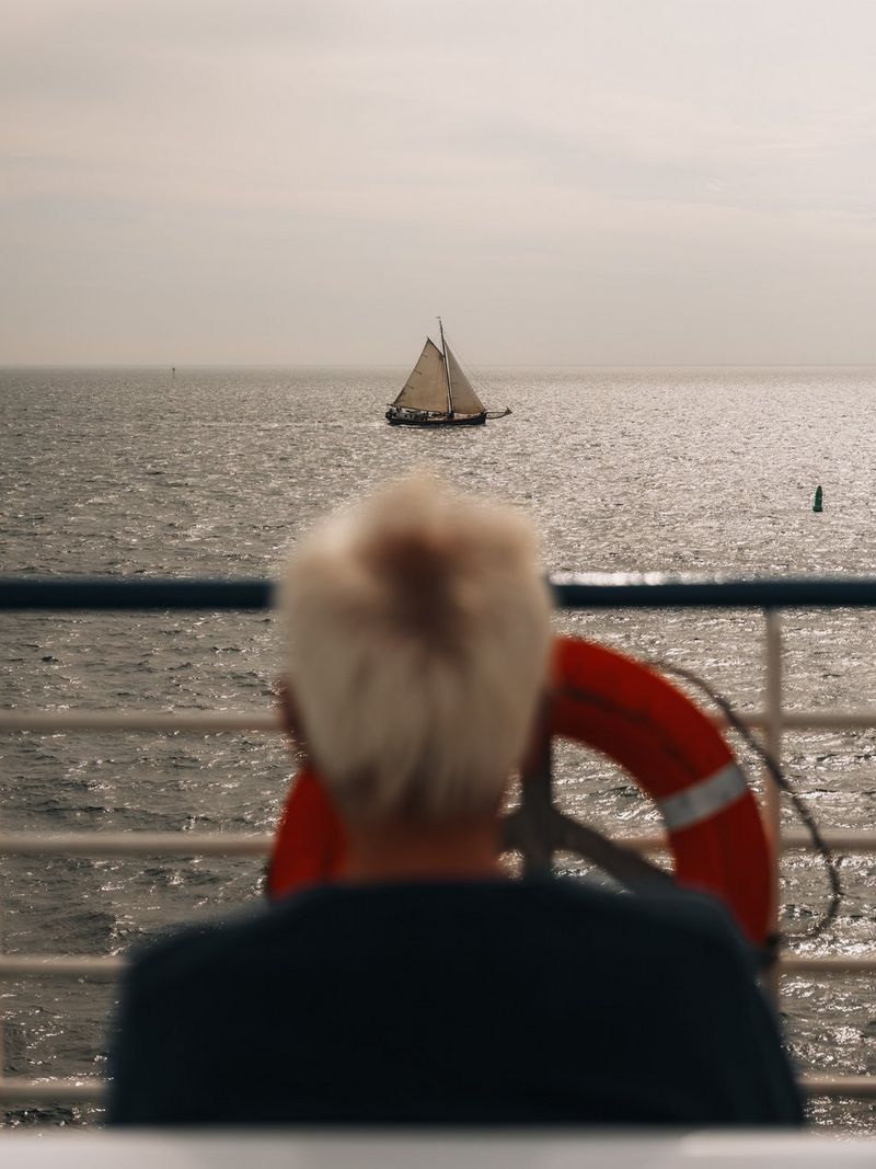 A man on the deck of a boat, blurred in the foreground, watches a sailing boat on the sea in the distance. Travel photo taken on a Canon EOS R5 Mark II.