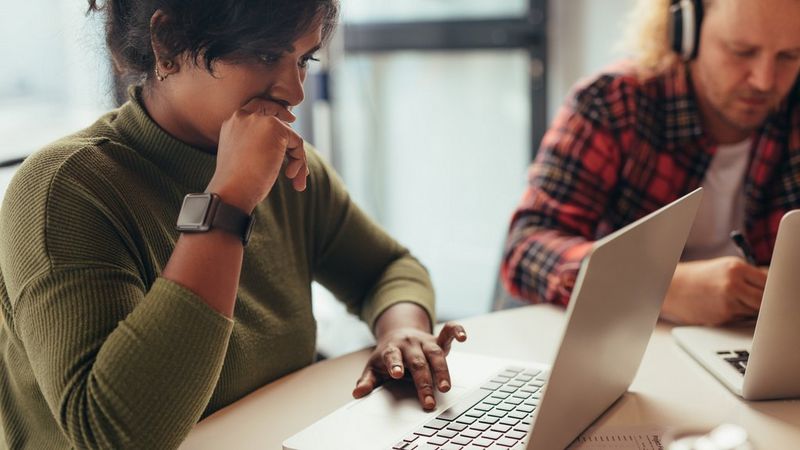 A woman sits at a table, looking at her laptop. Her hand is touching her chin, as though in deep concentration. In the background, another person with long, curly hair and headphones is also focused on a laptop.