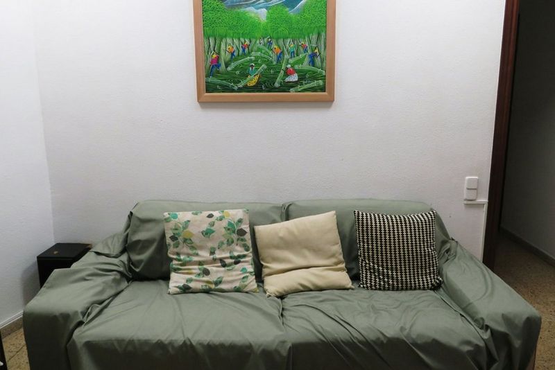 A light green sofa with three decorative pillows sits against a white wall beneath a framed painting of people in A green landscape.