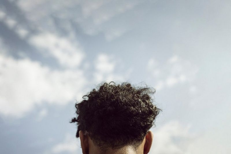 A close-up shot from behind shows a person's dark, curly hair against a backdrop of a bright, slightly cloudy sky.