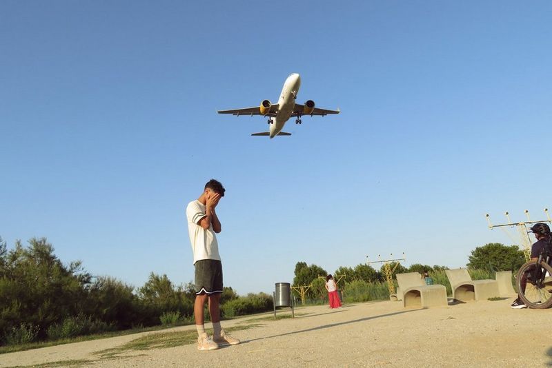 A person standing on a dirt path with their hands covering their ears as an aeroplane descends overhead in a clear, blue sky. There are other figures and greenery in the background.