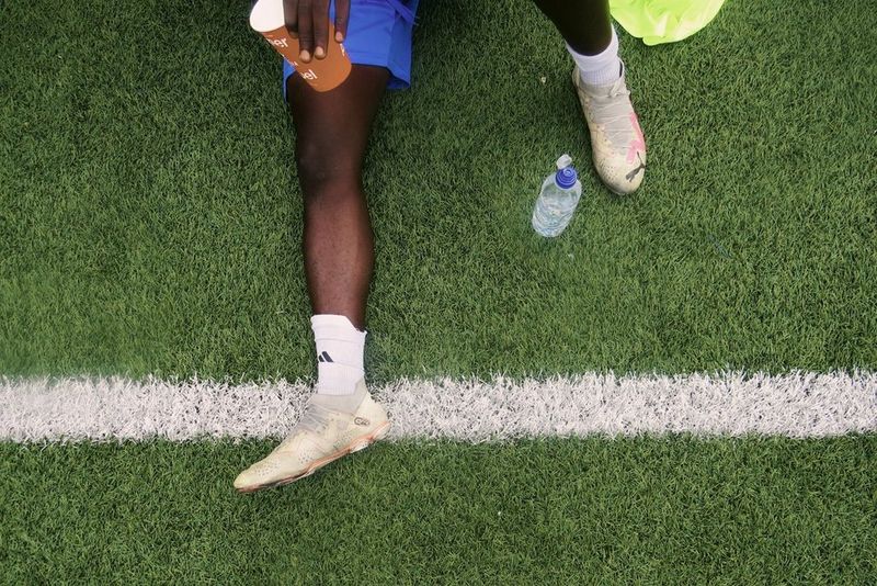 The legs of a person wearing blue shorts, white socks, and football boots. They are sitting on green turf with a white line, holding a water bottle, with a clear plastic water bottle nearby.