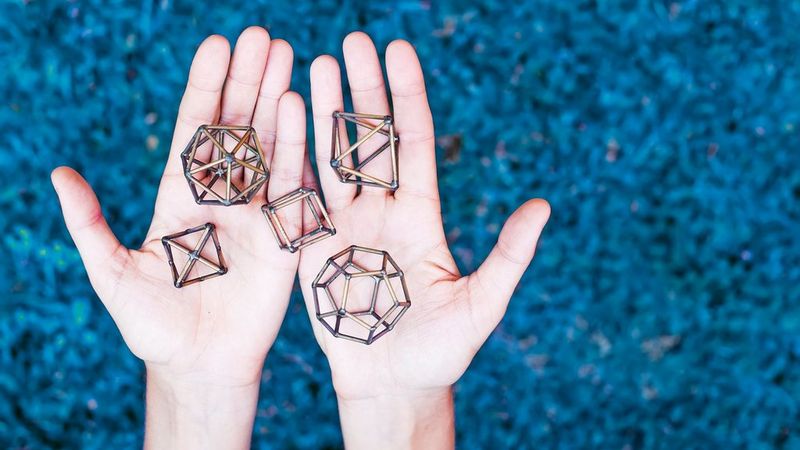 A pair of open hands, side by side. On them are five three dimensional geometric objects formed by connecting small rods. The background is a blurred uranium blue.