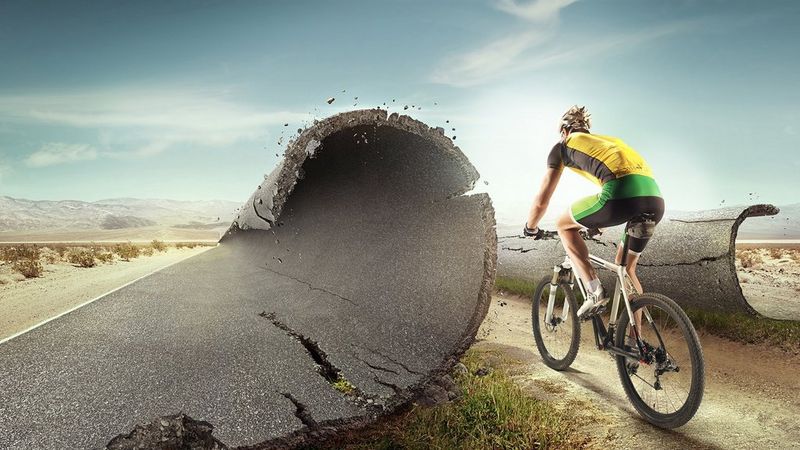 A concrete road is split in two, with both left and right sides curling and shedding debris as though to make way for a man who cycles through them, heading towards a desert landscape. He is wearing the lycra shorts, shirt and helmet of a professional cyclist.