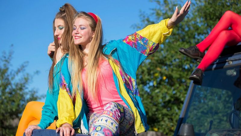 Two long-haired young women sit on the front of a jeep, smiling. They wear brightly coloured nineties-style shell suit jackets with bright leggings, one in bright acid orange and another in primary-coloured geometric shapes. They wear bright make up to match. The sky is bright clue and clear, and there are trees behind them. To their right, on the roof of the jeep is another person, but only their legs can be seen and are clad in bright red tights and black brogues.