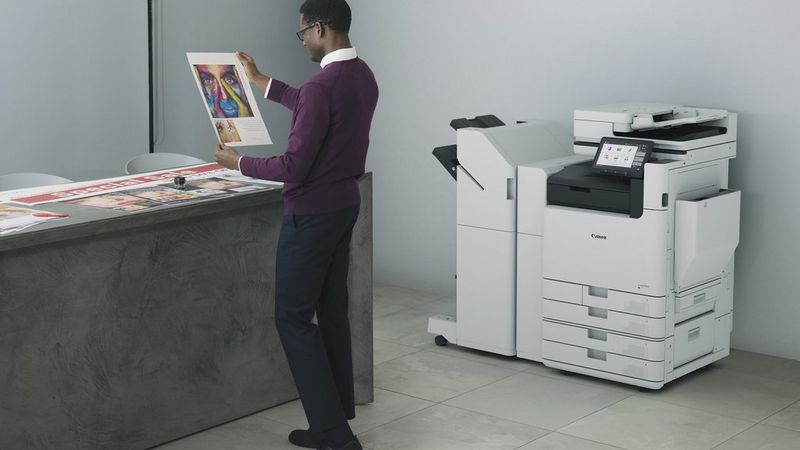 A man stands next to a Canon imageFORCE C7165 multifunction printer. He holds up a print and inspects it.