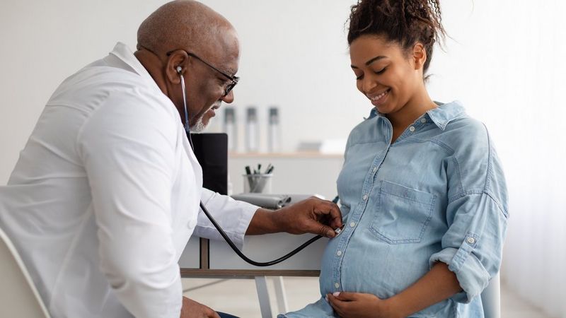A doctor in a white coat leans forward and presses a stethoscope against the belly of a smiling pregnant woman wearing a denim shirt.