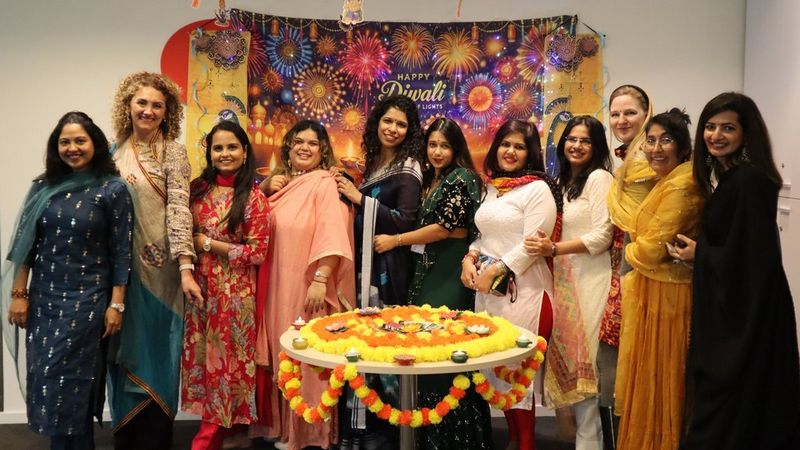 A group of women smiling together in front of a colourful ‘Happy Diwali’ banner. In front of them is a table decorated with a flower rangoli and small candles.