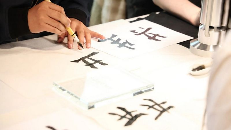 Close-up of a person's hand using a traditional brush and black ink to practice Japanese calligraphy on white paper.