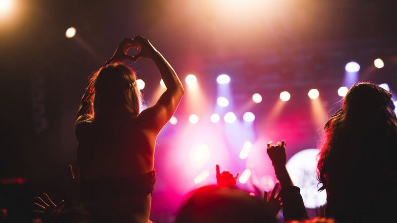 Shot taken from in the crowd at a concert. The stage lights shine down in the distance and hands are raised in the crowd. To the left, a young woman is on someone’s shoulders, her arms raised, making a heart shape with her hands.