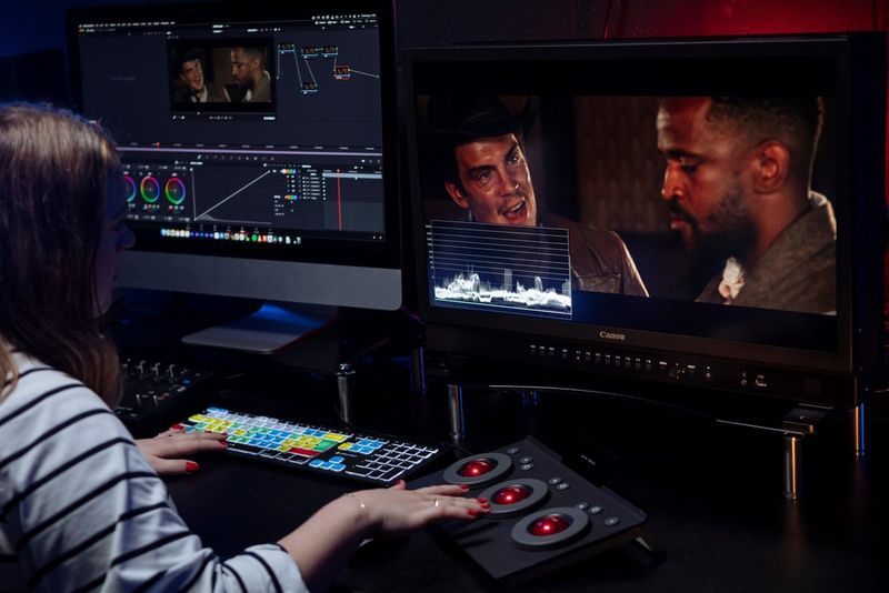 A woman sits at a desk on which is a Canon DP-V2730 reference monitor, speakers and video editing equipment.