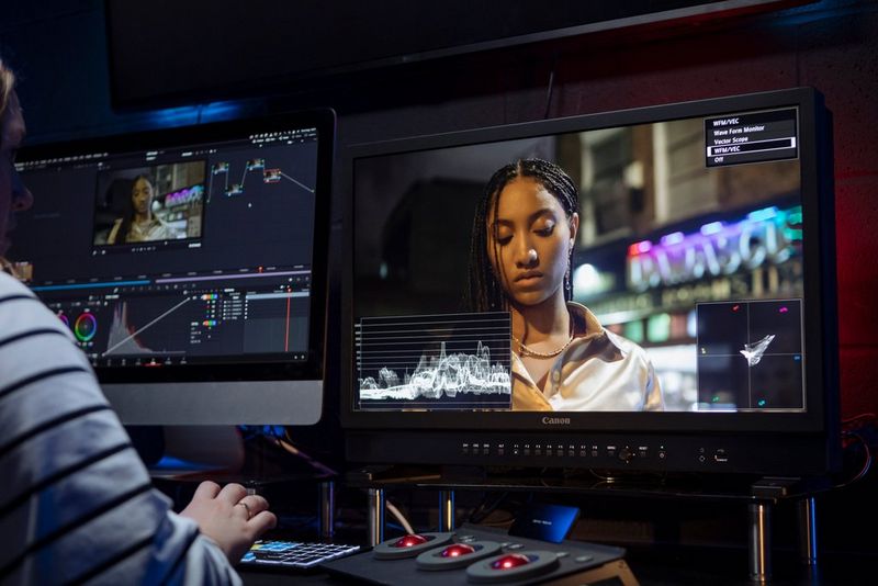 An editor sits in front of two large monitors, colour grading a video of a woman in an urban setting.