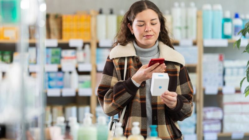 A young woman holds a boxed product in one hand and scans it with her smartphone. Shelves of personal care or pharmacy items are visible in the background.