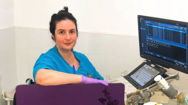 A dark haired woman in blue scrubs and purple latex gloves sits in front of a computer screen with assorted medical devices surrounding it. She has turned from the screen to pose for the camera.
