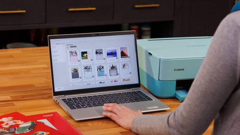 Een laptop staat open op een bureau met de Canon Easy-PhotoPrint-software op het scherm, Canon-papier aan de linkerkant en een Canon-printer aan de rechterkant.