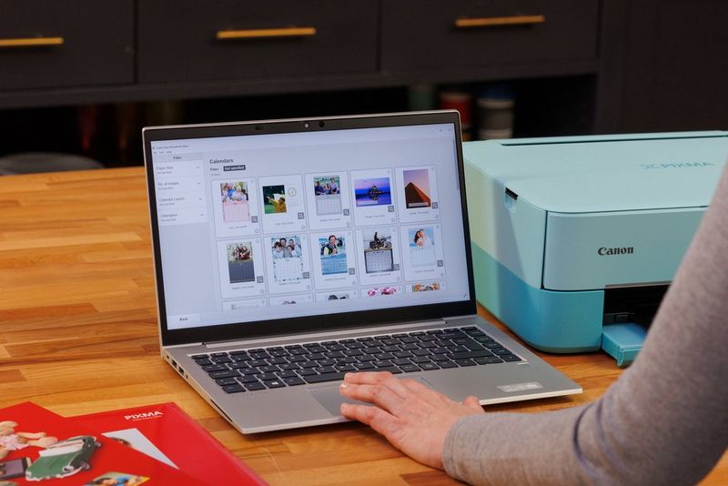 A laptop sits open on a desk with the Canon Easy-PhotoPrint software onscreen, Canon paper to the left and a Canon printer to the right. 