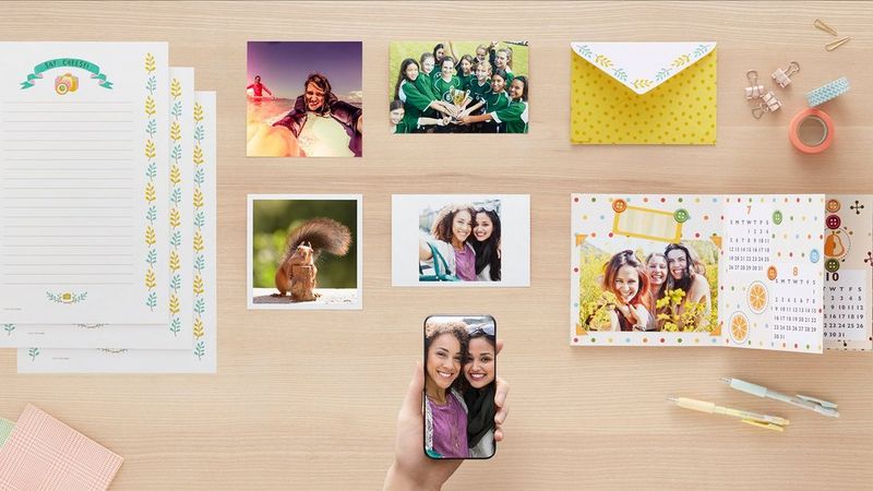 A flat lay image of a hand holding a smartphone, an envelope, calendar, note paper and printed pictures.